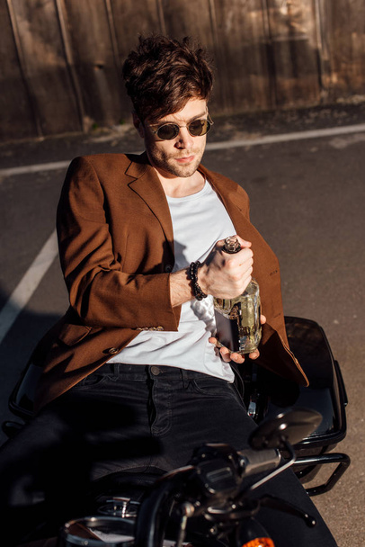homem bonito em terno marrom deitado na motocicleta e segurando garrafa de álcool
  - Foto, Imagem