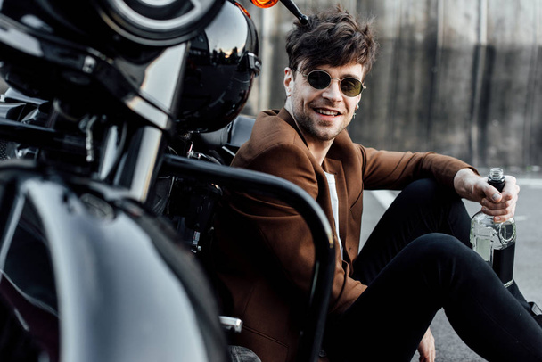 selective focus of handsome man sitting near motorcycle, holding bottle with alcohol and looking at camera - Photo, Image
