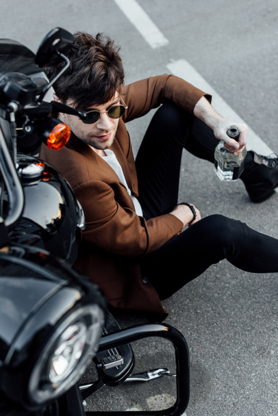 korkea kulma näkymä komea mies istuu gorund lähellä uutta moottoripyörää ja tilalla pullo
 - Valokuva, kuva