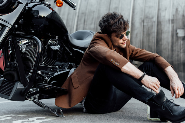 hombre con elegante chaqueta marrón sentado en el suelo cerca de la motocicleta, mirando hacia otro lado y sosteniendo la botella de alcohol
  - Foto, Imagen