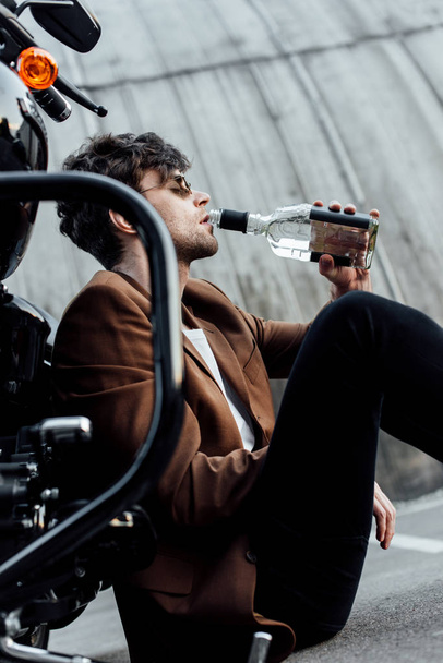 enfoque selectivo del hombre en gafas de sol y chaqueta marrón beber alcohol de la botella mientras está sentado en la motocicleta
 - Foto, Imagen