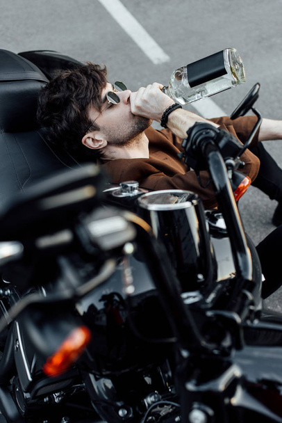 selective focus of man drinking alcohol while sitting on ground and leaning on motorcycle - Photo, Image