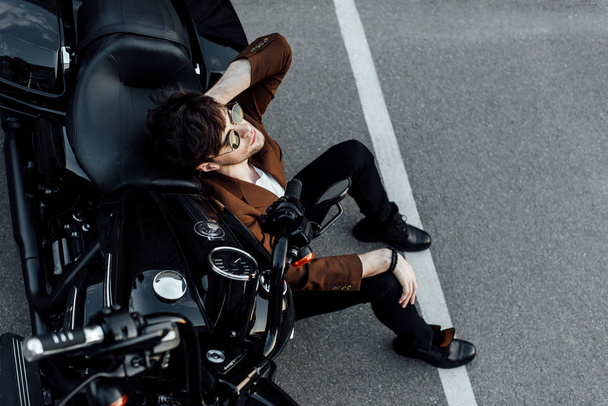 top view of man in sunglasses and suit sitting on ground near motorcycle - Foto, afbeelding