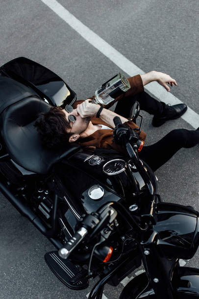Widok z góry człowieka w okulary przeciwsłoneczne i garnitur siedzi na ziemi w pobliżu motocykla i picia alkoholu z butelki  - Zdjęcie, obraz