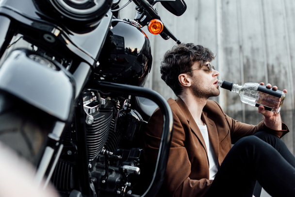 selective focus of man drinking alcohol while leaning on motorcycle and sitting on ground  - Foto, Imagem