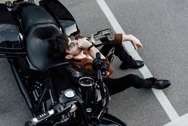 Blick von oben auf jungen Motorradfahrer, der auf dem Boden sitzt, sich auf Motorrad stützt und Alkohol trinkt - Foto, Bild