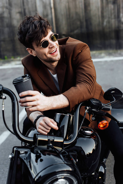 jonge man zittend op motorfiets, glimlachend, op zoek weg en het houden van papier kopje koffie  - Foto, afbeelding