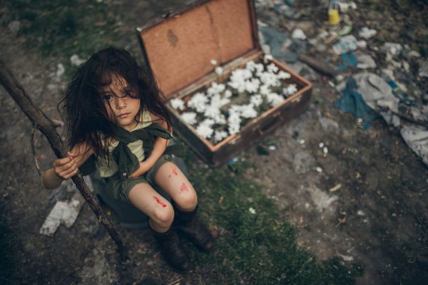 Bezdomna dziewczyna siedzi na śmieci obok walizki z kwiatami wewnątrz. - Zdjęcie, obraz