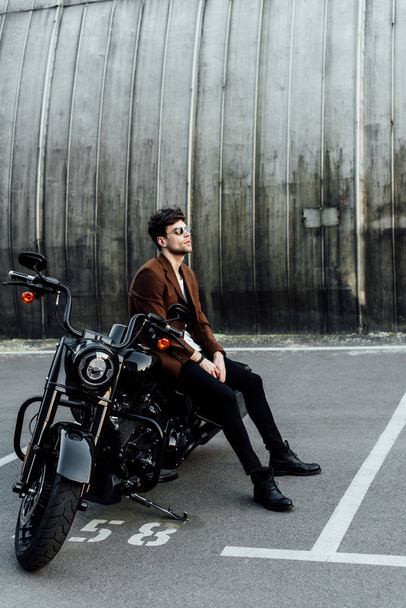 Ganzkörperansicht des Motorradfahrers ion braune Jacke sitzt auf Motorrad, ruht sich aus und schaut weg  - Foto, Bild
