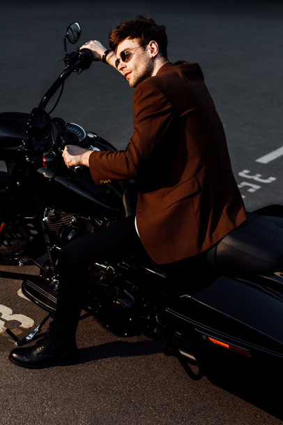 motorcyclist in brown jacket and sunglasses sitting on motorcycle and holding handlebars - Foto, imagen
