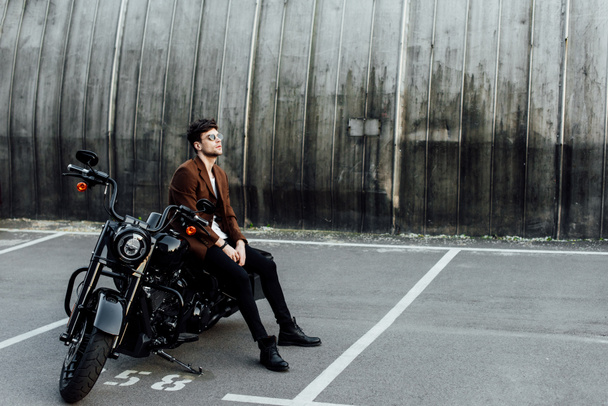 full length view of handsome man in jacket resting on black motorcycle and looking away - Zdjęcie, obraz