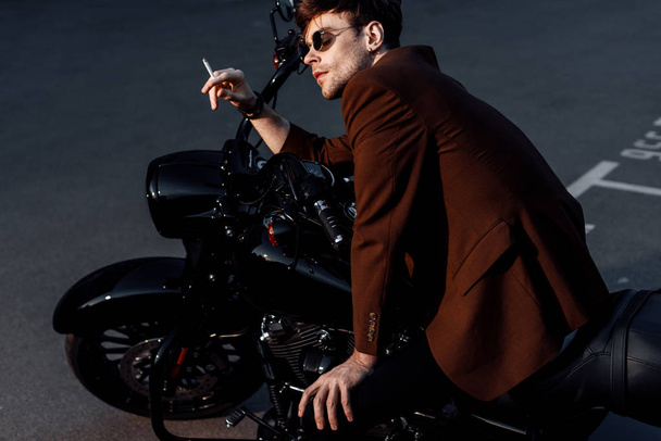 junger Mann in brauner Jacke sitzt auf schwarzem Motorrad und raucht Zigarette  - Foto, Bild