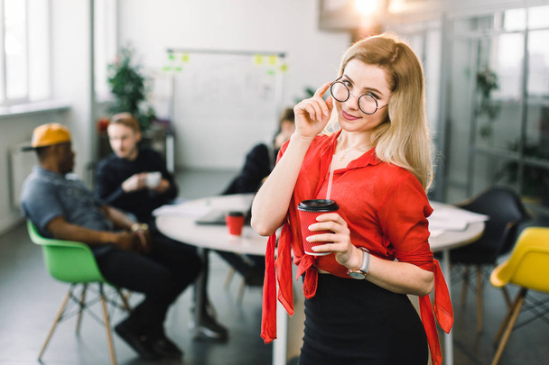 Cheerful young beautiful blond woman in glasses and red shirt standing at her working place on office with cup of coffee. Multiracial young creative people in modern office. - Photo, Image