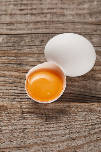gebroken eierschaal met gele dooier in de buurt van ei op houten tafel - Foto, afbeelding