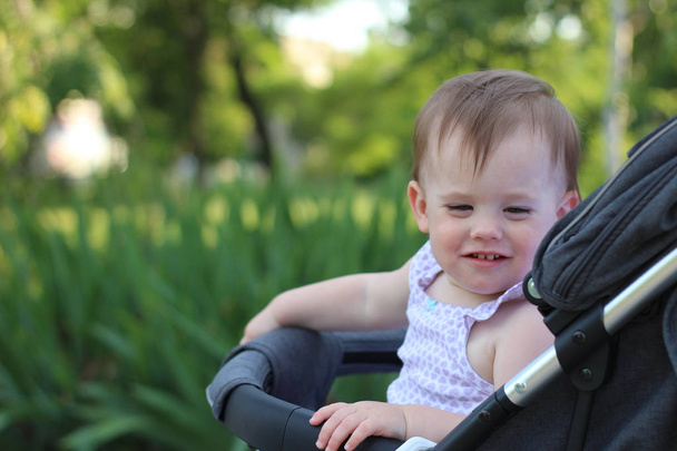 kleines, schönes, lächelndes, niedliches rothaariges Baby im Kinderwagen draußen im ärmellosen Hemd, das nach unten schaut - Foto, Bild