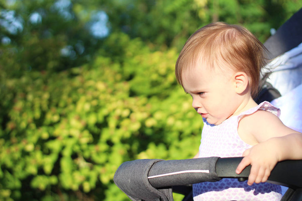 little, beautiful, smiling, cute redhead baby in a pram out-of-doors in a sleeveless shirt looking down - Photo, Image