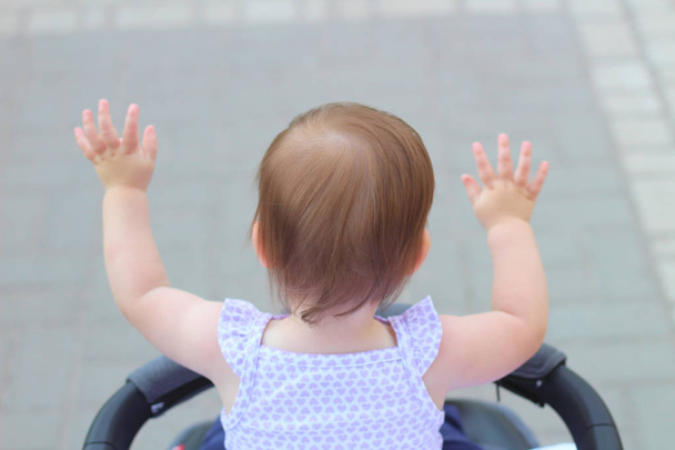 pequeno, lindo, sorrindo, bebê ruiva bonito em uma camisa sem mangas em um carrinho fora de portas levanta as mãos para cima olhando para a frente
 - Foto, Imagem