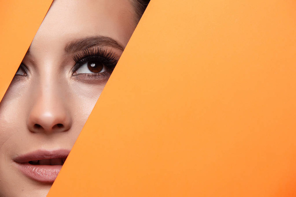 A girl with beautiful curry bright beautiful eyes with brown shadows and expressive eyebrows looks into the hole of colored paper.Fashion, beauty, make-up, cosmetics, make-up artist, beauty salon,business card. - Foto, Imagem