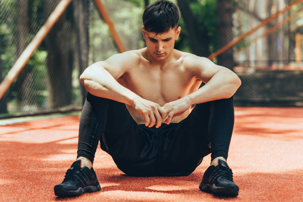 Athlete male taking a rest after training session outdoors. Healthy and fit man sitting at pavement sportsground and realxing after workout exercises. People and sport concept - Φωτογραφία, εικόνα
