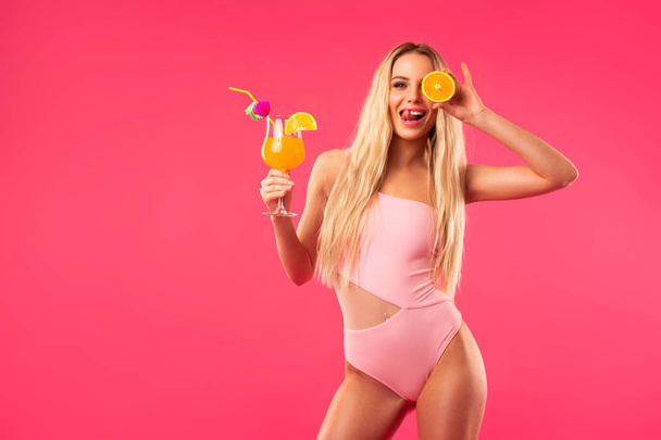 blondes Mädchen im Badeanzug mit einem Cocktail und einer halben Orange in den Händen  - Foto, Bild