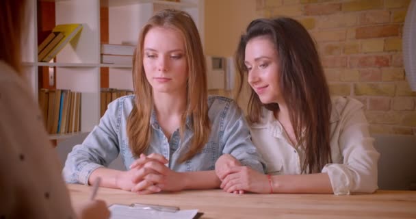 Closeup shoot of young beautiful lesbian couple talking to female realtor about purchase of an apartment signing a document and getting the keys sitting by the table indoors - Imágenes, Vídeo
