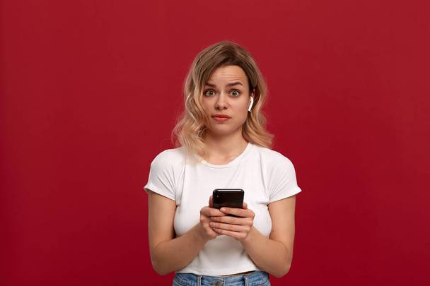 Πορτρέτο ενός κοριτσιού με σγουρά ξανθά μαλλιά σε ένα λευκό μπλουζάκι σε κόκκινο φόντο. Δυσαρεστημένο μοντέλο σε ασύρματα ακουστικά κοιτάζει τη φωτογραφική μηχανή και κρατά το κινητό τηλέφωνο.  - Φωτογραφία, εικόνα