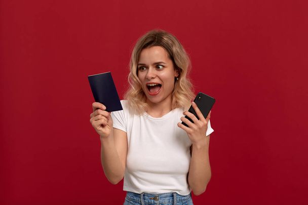 Portrait of a girl with curly blond hair in a white t-shirt on a red background. Happy model looks at the screen of a mobile phone and holds passport. - Foto, immagini