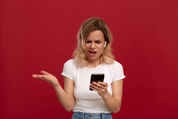 Porträt eines Mädchens mit blonden Locken in einem weißen T-Shirt auf rotem Hintergrund. Modell mit unzufriedenem Blick im drahtlosen Headset blickt auf den Bildschirm eines Mobiltelefons. - Foto, Bild