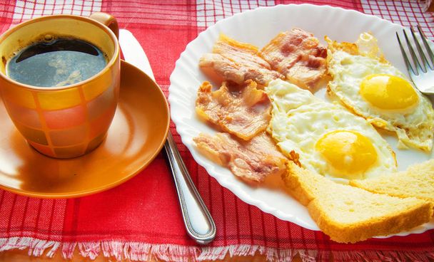 Finom reggeli - egy csésze kávé, egy tányér sült tojás, szalonna és pirítós, mellett az evőeszközök piros kockás szalvéta - Fotó, kép