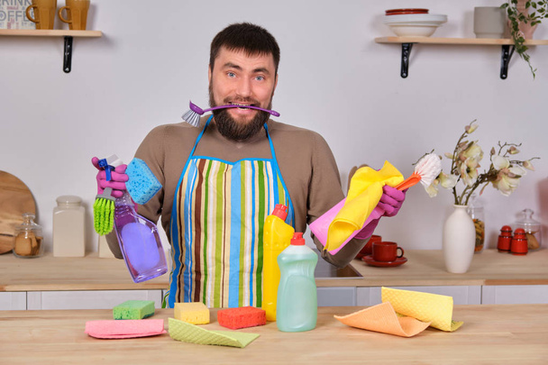 Запутавшийся молодой красивый бородатый мужчина на кухне, понятия не имеет, что делать со всеми его моющими средствами, щетками, спреями
 - Фото, изображение