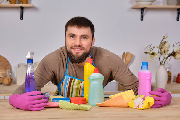 Joven hombre barbudo guapo en la cocina, muestra todo su personal de limpieza detergentes, cepillos, aerosoles. Cree que está listo para una verdadera limpieza.
 - Foto, imagen