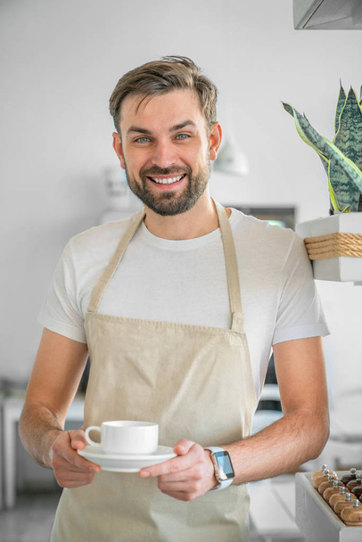 Vertical shot of happy coffee shop owner posing with a coffee cup in hand. Closeup of barista standing at the entrance of coffee shop holding a coffee cup. - Foto, Imagen