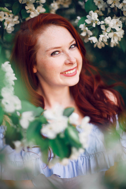 Model with red hair posing in a blooming apple tree. - Foto, afbeelding