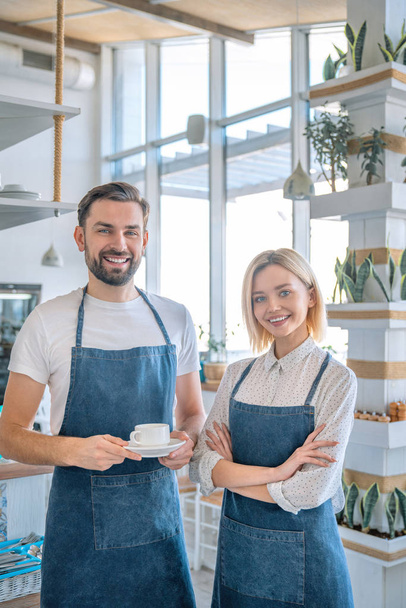 Happy coffee shop owner couple standing inside their shop. Man and woman baristas standing inside their cafe wearing apron with the man holding coffee cup - Foto, Imagem