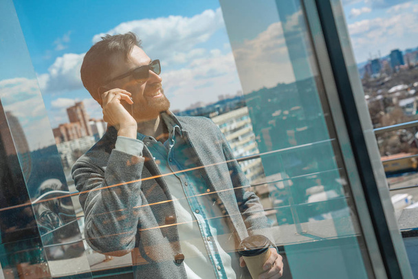 Side portrait of a smiling man in sunglasses talking on mobile phone with cup of coffee standing on a roof top - Photo, image