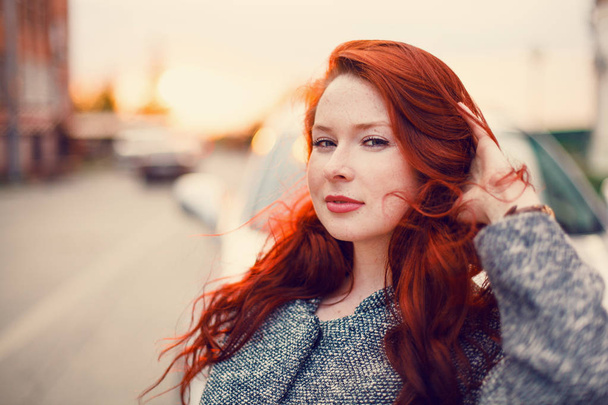 Red-haired model in a coat posing against the backdrop of a sunset on the street. - Fotó, kép