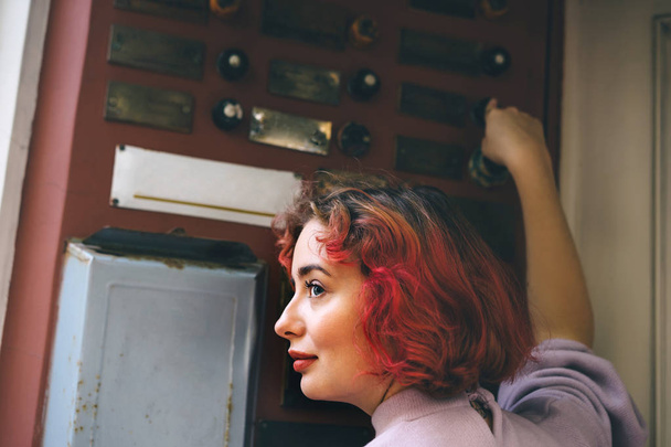 Beautiful young woman with orange hair ringing the door of an apartment  - Foto, afbeelding