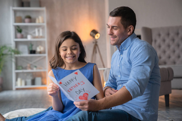 Happy Father's Day! Caucasian young man at home with his little cute girl look at gift postcard - Photo, Image