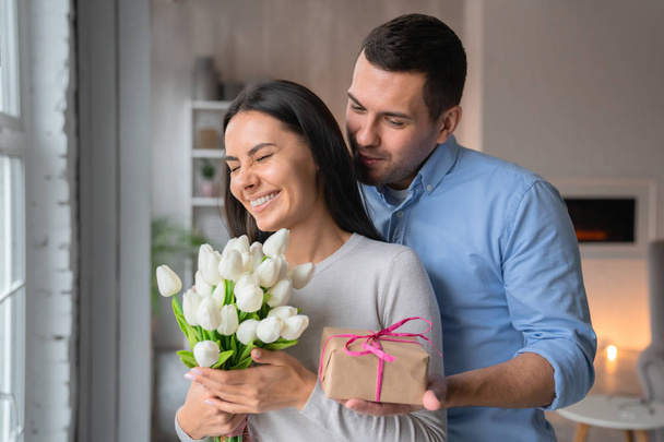 Close up shot of cheerful young caucasian man that giving present box to his wife with look on her. Beautiful woman excited with flowers in hands in home interior , happy couple - Foto, imagen