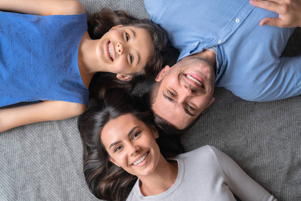 Happy family concept. Top view of happy family of three bonding to each other and smiling while lying on the floor - Zdjęcie, obraz
