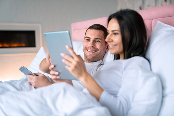 Cheerful woman showing something in tablet to her husband. Smiling man lying on bed with his wife holding mobile phone in his hands with focus on man - Zdjęcie, obraz