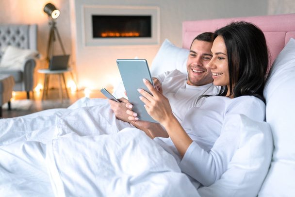 Smiling woman showing something in tablet to her husband. Smiling man lying on bed with his wife holding mobile phone in his hands - Foto, Imagem