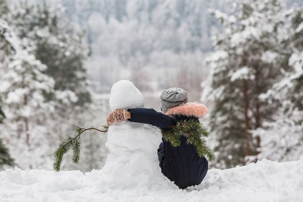 冬の雪の公園で雪だるまとお互いを抱きしめ合う少女の背面図. - 写真・画像