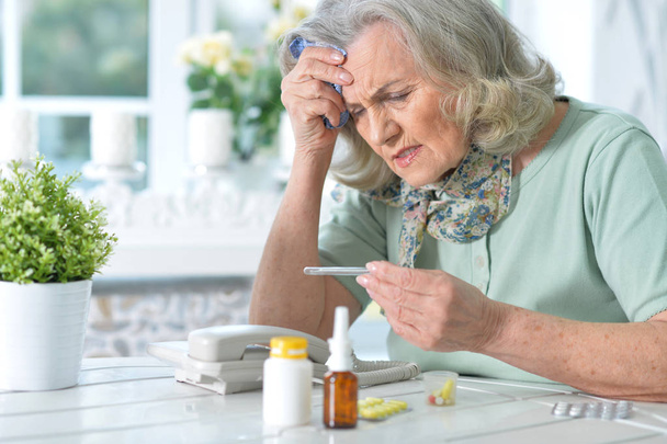Mujer mayor enferma sentada en la mesa en casa con termómetro
  - Foto, Imagen