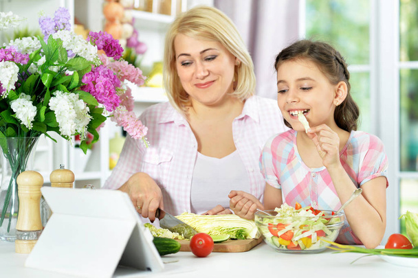 Cute little girl with her mother cooking together at kitchen table with tablet on it  - Foto, imagen