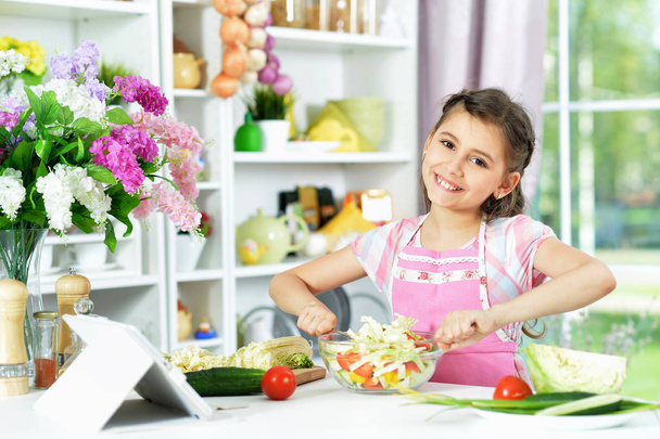 Cute little girl preparing fresh salad on kitchen table with tablet at home - Zdjęcie, obraz