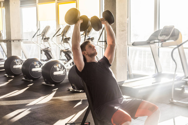 concentrated young adult handsome athlete working out in gym while sitting on bench and holding two dumbbell with raised arms and doing exercises for biceps - Foto, Bild