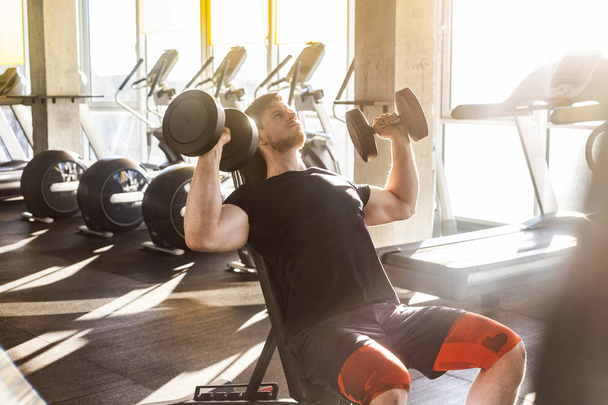 concentrated young adult handsome athlete working out in gym while sitting on bench and holding two dumbbell with raised arms and doing exercises for biceps - Foto, immagini
