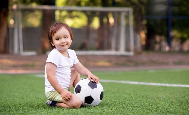 Little toddler boy in sports uniform sitting with soccer ball at football field outdoors in summer day. Goalposts (soccer nets) in background. Kid sports and active childhood concept. Room for copy text - Fotoğraf, Görsel