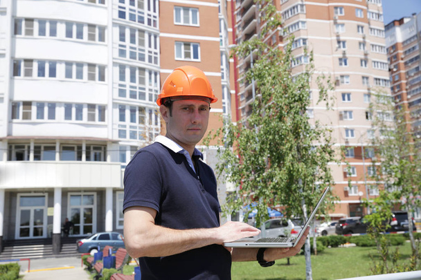 Retrato de un joven guapo con un casco protector con un portátil en las manos frente a un edificio alto. Construcción, arquitectura y negocios
. - Foto, imagen